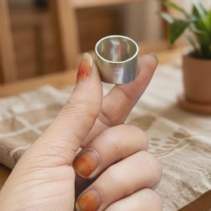 May include: A wide, polished silver-toned band ring with a smooth, reflective surface and a cylindrical shape. The ring features a consistent width throughout its circumference, showcasing a clean, metallic finish without any additional engravings or stones.