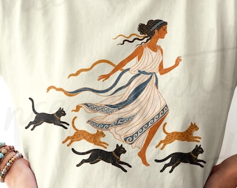 Pode incluir: Uma t-shirt de manga curta na cor marfim com uma ilustração gráfica de uma figura com uma peça drapeada branca e azul a correr ao lado de quatro gatos. Os gatos são pretos e castanhos e a figura tem cabelo escuro preso atrás.