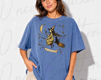 Pode incluir: Uma t-shirt azul de manga curta com um gráfico de uma bruxa com um gato preto numa vassoura, uma lua crescente e uma faixa com o texto "Meet me under the Harvest Moon" rodeado por uma borda de videira com folhas e estrelas.