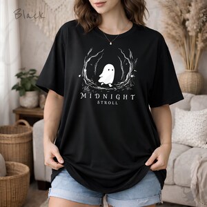 Op de afbeelding: Een zwart t-shirt met korte mouwen en een witte afbeelding van een klein spookje dat door een gestileerd bos loopt. Onder het spookje staat de tekst MIDNIGHT STROLL in witte hoofdletters. Het shirt heeft een ronde halslijn.
