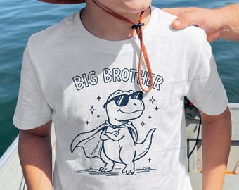 Pode incluir: Uma t-shirt branca de manga curta com um gráfico de linhas azul-escuro de um dinossauro a usar óculos de sol e uma capa. O texto "BIG BROTHER" está impresso num arco acima do dinossauro, rodeado por pequenos ícones de estrelas.