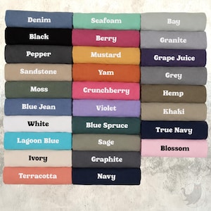 Op de afbeelding: Een verzameling opgevouwen t-shirts gerangschikt in een raster, elk gelabeld met kleurnamen waaronder Denim, Black, Pepper, Sandstone, Moss, Blue Jean, White, Lagoon Blue, Ivory, Terracotta, Seafoam, Berry, Mustard, Yam, Crunchberry, Violet, Blue Spruce, Sage, Graphite, Navy, Bay, Granite, Grape Juice, Grey, Hemp, Khaki, True Navy en Blossom.