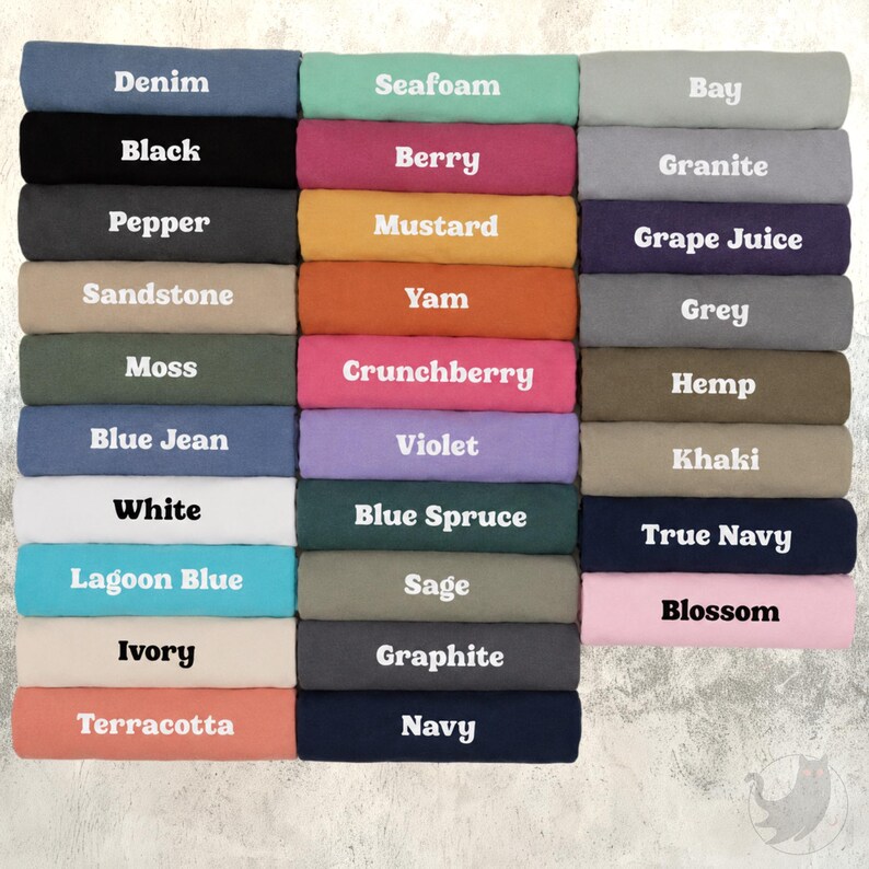Op de afbeelding: Een verzameling opgevouwen t-shirts gerangschikt in een raster, elk gelabeld met kleurnamen waaronder Denim, Black, Pepper, Sandstone, Moss, Blue Jean, White, Lagoon Blue, Ivory, Terracotta, Seafoam, Berry, Mustard, Yam, Crunchberry, Violet, Blue Spruce, Sage, Graphite, Navy, Bay, Granite, Grape Juice, Grey, Hemp, Khaki, True Navy en Blossom.