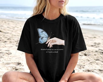Pode incluir: Uma t-shirt preta de manga curta com um gráfico de uma borboleta azul-clara pousada numa mão. Abaixo do gráfico, o texto diz: Kindred spirits are not so scarce as I used to think. A t-shirt tem um decote redondo e um corte descontraído.