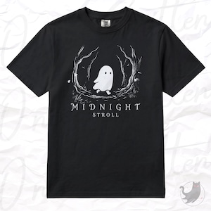 Op de afbeelding: Een zwart t-shirt met korte mouwen en een witte afbeelding van een klein spookje dat door een gestileerd bos loopt. Onder de illustratie staat de tekst MIDNIGHT STROLL in witte hoofdletters. Het shirt heeft een ronde hals en een standaard pasvorm.