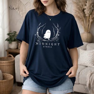 Op de afbeelding: Een marineblauw t-shirt met korte mouwen en een witte afbeelding van een klein spookje dat door een bos met kale bomen loopt. Onder het spookje staat de tekst MIDNIGHT STROLL in een strak, wit lettertype. Het shirt heeft een ronde hals.