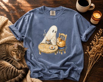 Pode incluir: Uma t-shirt azul de ganga com um gráfico de um fantasma branco sentado a uma mesa redonda de madeira com uma chávena de café, enquanto um gato laranja está sentado numa cadeira de madeira por perto. O fantasma segura uma chávena com vapor.
