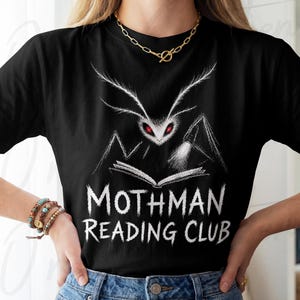 May include: A black short-sleeved t-shirt featuring a white graphic of a moth-like creature with glowing red eyes reading an open book with a flashlight. The text "MOTHMAN READING CLUB" is printed in a distressed white font below the illustration.
