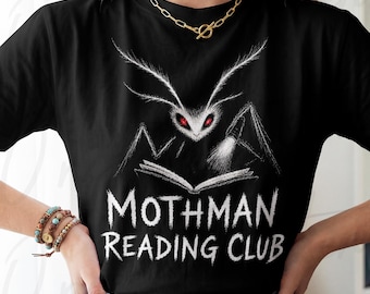 Pode incluir: Uma t-shirt preta de manga curta com um gráfico branco de uma criatura semelhante a uma traça com olhos vermelhos brilhantes a ler um livro aberto com uma lanterna. O texto "MOTHMAN READING CLUB" está impresso numa fonte branca.