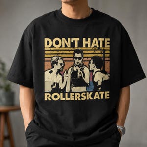 May include: A black short-sleeved t-shirt featuring a graphic print with the text "DON'T HATE ROLLERSKATE" in a bold font. The design includes a retro-style sunset background and a stylized illustration of three figures in a vintage aesthetic.