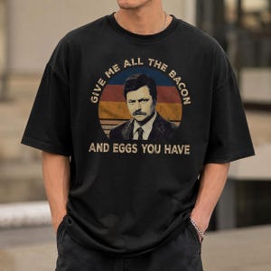 May include: A black short-sleeved t-shirt featuring a circular graphic with a sunset-inspired background of blue, orange, and red stripes. The graphic displays a portrait of a man with a mustache and the text GIVE ME ALL THE BACON AND EGGS YOU HAVE.