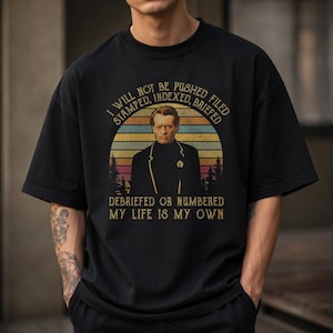May include: A black short-sleeve t-shirt featuring a graphic of a man in a dark blazer. The text reads: I WILL NOT BE PUSHED FILED STAMPED, INDEXED, BRIEFED DEBRIEFED OR NUMBERED MY LIFE IS MY OWN. The design includes a sunset background.