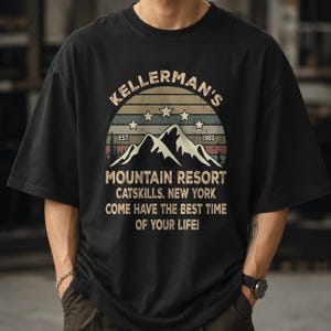 May include: A black short-sleeve t-shirt featuring a graphic print of a mountain range under a sunset arc. The text reads: KELLERMAN'S MOUNTAIN RESORT EST. 1963 CATSKILLS, NEW YORK COME HAVE THE BEST TIME OF YOUR LIFE! Five stars are arranged in an arc.