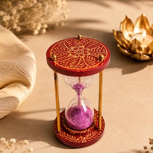 Puede incluir: Un reloj de arena con un bulbo de vidrio transparente que contiene arena morada. Las bases circulares de madera superior e inferior están pintadas de rojo intenso con intrincados patrones florales dorados. Cuatro varillas metálicas doradas unen ambos extremos.