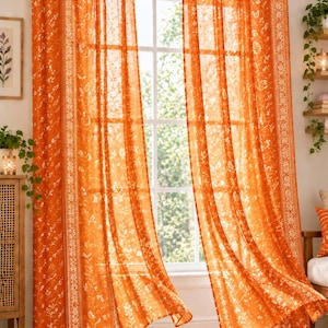 Könnte beinhalten: Ein Paar leuchtend orangefarbene Vorhänge mit einem zarten weißen Blumenmuster und einem vertikalen Bordürendesign. Die Vorhänge sind mit Stoffschlaufen an einer Metallstange befestigt, wodurch Licht durch das halbtransparente Material fällt.