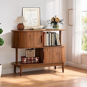 May include: A walnut wood console table with rounded ends, featuring two sliding tambour doors, an open top shelf, and a lower shelf. The unit is supported by four tapered legs and includes a small woven basket and a plaid storage box inside.
