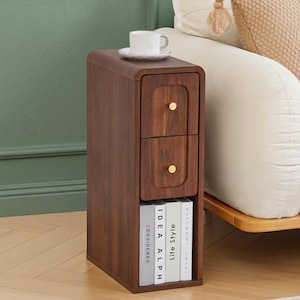 May include: A narrow, dark wood side table featuring two stacked drawers with small, round gold-toned knobs. The top surface holds a white ceramic mug on a matching saucer. Below the drawers is an open shelf containing three books standing upright.
