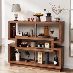 May include: A multi-tiered wooden shelving unit with a walnut finish, featuring an open geometric design supported by silver metal poles. The shelves are filled with various kitchen items, including a lamp, fruit bowl, coffee grinder, glassware, and food.