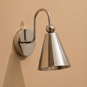 May include: A polished chrome wall sconce featuring a circular mounting plate, a curved arm, and a conical shade. The fixture includes a spherical accent piece connecting the arm to the shade, all finished in a reflective, metallic silver tone.