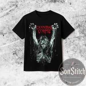 Puede incluir: Una camiseta negra de manga corta con un estampado gráfico de una figura esquelética con pelo largo y brazos levantados. El texto CANNIBAL CORPSE está impreso en rojo sobre la figura, representada en tonos blancos y azul claro con efectos de goteo.