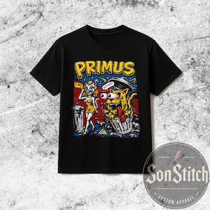 Może przedstawiać: Czarny t-shirt z krótkim rękawem z nadrukiem graficznym z napisem PRIMUS u góry. Ilustracja przedstawia postać z kreskówki w żółtej sukience stojącą obok kosza na śmieci oraz żółtego kota z wyłupiastymi oczami wewnątrz kubła na śmieci.