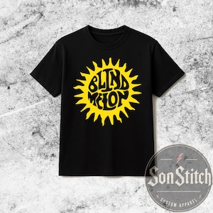 Op de afbeelding: Een zwart T-shirt met korte mouwen en een grote, felgele afbeelding van een zon. In het midden van de zon staat de tekst "BLIND MELON" in een gestileerd, golvend lettertype. Het shirt heeft een ronde hals en een rechte zoom aan de onderkant.