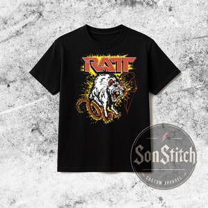 May include: A black short-sleeve t-shirt featuring a graphic print of a white rat with red eyes and bared teeth. Above the rat, the word RATT is printed in red block letters against a yellow splatter background. A circular logo reads SonStitch.