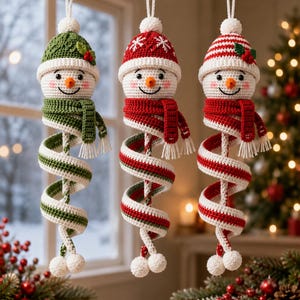 May include: Three crocheted snowman ornaments hanging vertically. Each features a white head with a carrot nose, rosy cheeks, and a unique hat. They wear long, spiralling scarves in green and white, red and white, or striped patterns with pom-poms.