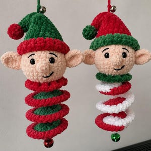 Puede incluir: Dos adornos de elfo hechos a mano en ganchillo con cabezas de felpa, gorros puntiagudos, orejas grandes y caras sonrientes. El elfo izquierdo tiene un gorro verde y cuerpo en espiral rojo y verde, el derecho un gorro rojo y cuerpo rojo y blanco.