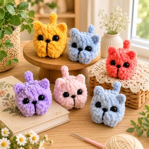 May include: Six small, round, crocheted cat figures made of soft, textured yarn in yellow, light blue, coral, purple, light pink, and pale blue. Each cat features two pointed ears, a small upright tail, and two large, round, black plastic eyes.