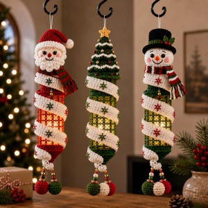 May include: Three hanging cylindrical Christmas decorations featuring crocheted characters: a Santa Claus, a green Christmas tree, and a snowman. Each ornament is wrapped in a white crocheted ribbon with red and green snowflake embroidery.