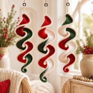 Puede incluir: Tres adornos colgantes en espiral hechos de hilo de ganchillo. Cada adorno presenta un diseño retorcido y ondulado con bandas alternas de color rojo, verde oscuro y blanco, acabado con un borde de ganchillo crema y una pequeña anilla negra.