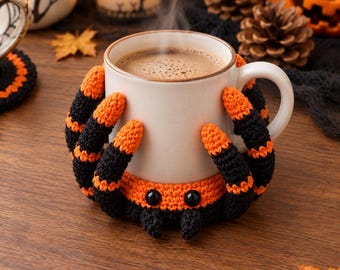 Puede incluir: Una taza de cerámica blanca llena de una bebida marrón humeante, rodeada por una funda de ganchillo con forma de araña. La funda presenta patas a rayas negras y naranjas, un cuerpo negro y naranja y dos ojos negros brillantes en la base.