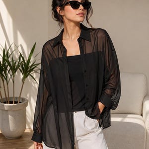 May include: A sheer black button-down shirt with a classic collar and long sleeves, worn over a solid black camisole. The shirt features a lightweight, translucent fabric, a front button placket, and cuffed sleeves, paired with light-colored trousers.