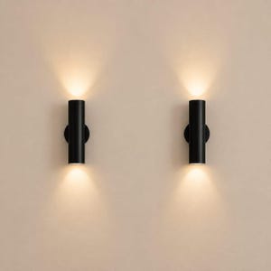 Peut inclure: Deux appliques murales cylindriques noires montées verticalement. Chaque luminaire possède une base circulaire et émet un faisceau lumineux chaud et concentré par les ouvertures supérieure et inférieure, créant une lueur symétrique.