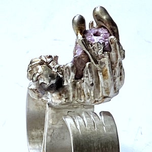 Könnte beinhalten: Ein silberner Doppelring mit strukturierter Oberfläche. Der Ring zeigt ein Reliefdesign aus zwei Händen, die einen großen, violetten Edelstein halten.