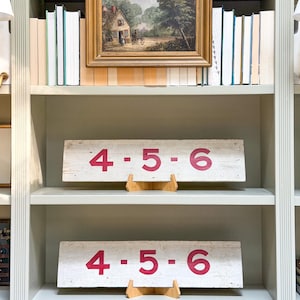 May include: Two rectangular white wooden signs, each featuring the red painted numbers 4, 5, and 6 separated by dots. Each sign is displayed on a small wooden stand, positioned on separate shelves of a light-colored bookcase with books.