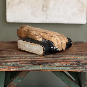 May include: A carved wooden sculpture of a human hand resting flat on top of a rectangular book. The hand features a natural light brown wood grain finish, while the book has a dark black cover with visible white pages along the side edge.