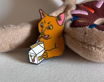 May include: A gold and brown enamel pin featuring a cartoon cat drinking from a milk carton. The milk carton is white with a cow graphic and the word "MILK". The cat has pink ears and is holding the carton with a straw.