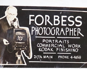 Può includere: Foto in bianco e nero di un uomo che tiene una grande macchina fotografica. Il testo "FORBESS PHOTOGRAPHER" è sopra il testo "PORTRAITS COMMERCIAL WORK KODAK FINISHING 217 1/2 MAIN PHONE 4-4659".