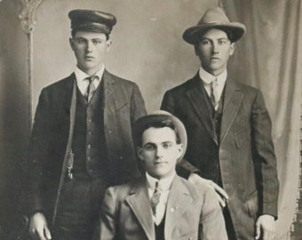 Può includere: Tre uomini in abito e cappello posano per un ritratto. L'uomo al centro è seduto, mentre gli uomini ai lati sono in piedi. Tutti e tre gli uomini indossano abiti e cravatte. L'uomo a sinistra indossa un berretto, mentre gli uomini a destra indossano cappelli.