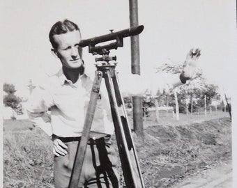 Può includere: Una fotografia in bianco e nero di una persona in piedi accanto a un treppiede con uno strumento di topografia. La persona sta guardando attraverso lo strumento.
