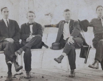 Può includere: Quattro uomini in abito e cappello sono seduti su una panchina. Guardano tutti la fotocamera. Gli uomini indossano cravatte e papillon. La panchina è in legno e ha una superficie bianca.