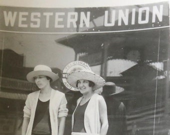 Könnte beinhalten: Zwei Frauen in Hüten und Strickjacken stehen vor einem Schaufenster mit dem Text "WESTERN UNION" darüber. Die Frauen tragen kurzärmelige Strickjacken und kurze Röcke.