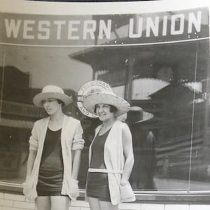 Puede incluir: Dos mujeres con sombreros y cárdigans se paran frente a una vidriera con el texto "WESTERN UNION" arriba. Las mujeres llevan cárdigans de manga corta y faldas cortas.