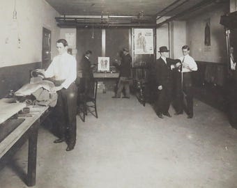 Può includere: Foto in bianco e nero di un gruppo di uomini in un laboratorio. Gli uomini indossano abiti e cappelli. C'è un tavolo al centro della stanza con un grande pezzo di stoffa sopra. Gli uomini sembrano lavorare sulla stoffa.