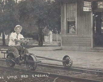 Può includere: Un uomo con un cappello bianco e una camicia bianca guida un piccolo veicolo rosso a propulsione ferroviaria. Il veicolo ha un solo sedile e un telaio lungo e stretto. L'uomo sorride e sembra godersi il giro. Il veicolo è etichettato "The Little Red Special".
