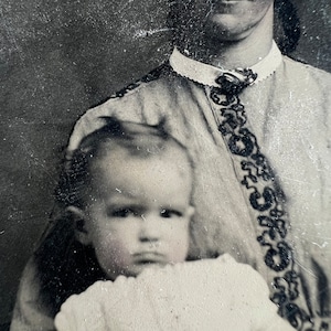 May include: A black and white vintage photograph of a woman holding a young child. The woman is wearing a high-collared blouse with a decorative trim. The child is looking directly at the camera.