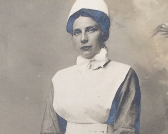 May include: A black and white photo of a woman wearing a white nurse's uniform with a white cap and a white bow tie. She is looking directly at the camera.