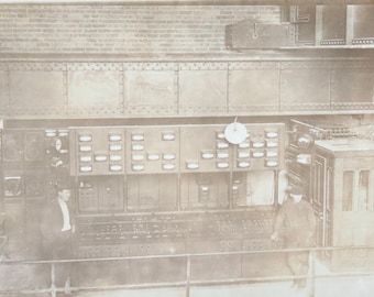 Può includere: Una fotografia in bianco e nero di un pannello di controllo vintage con numerosi interruttori e indicatori. Il pannello è in metallo e ha un aspetto usurato e industriale. Due persone sono in piedi davanti al pannello, una su ogni lato.